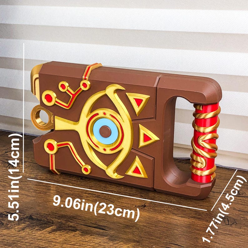 Pode incluir: Um adere&ccedil;o castanho com detalhes dourados e vermelhos, com uma pega e padr&otilde;es geom&eacute;tricos. O adere&ccedil;o mede 23 cm de comprimento, 14 cm de altura e 4,5 cm de largura. O design inclui um s&iacute;mbolo central em forma de olho.