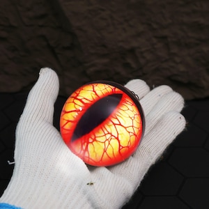 May include: A spherical ornament resembling a glowing eye, featuring a black pupil and a red and yellow iris with red vein-like patterns. The object is held in a white-gloved hand. The background is a dark, textured surface.