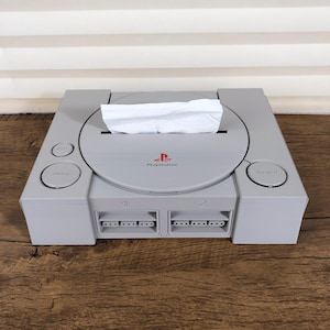 Op de afbeelding: Een grijze PlayStation 1 console omgebouwd tot een tissuebox. De console heeft het PlayStation-logo en het woord "PlayStation". Witte tissues zijn zichtbaar in de schijfstationsleuf. De console staat op een houten oppervlak.