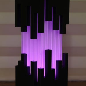 Puede incluir: Lámpara abstracta con diseño geométrico negro. Bloques rectangulares negros verticales dispuestos sobre una fuente de luz púrpura translúcida y brillante. El diseño es moderno y artístico, creando un efecto visual llamativo.