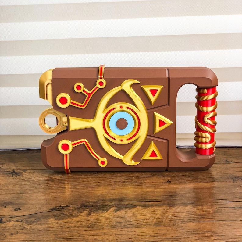 Pode incluir: Um adere&ccedil;o castanho com detalhes dourados, vermelhos e azuis. O adere&ccedil;o apresenta uma pega envolta em vermelho e dourado, um design circular com um centro azul e uma s&eacute;rie de formas geom&eacute;tricas vermelhas e douradas. O adere&ccedil;o tem uma forma retangular com uma pega.