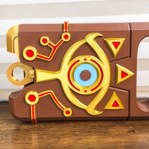 Pode incluir: Um objeto decorativo castanho, dourado, vermelho e azul com formas geom&eacute;tricas. O objeto apresenta um design central em forma de olho, tri&acirc;ngulos vermelhos e uma abertura circular. O design tem um tema futurista ou de fantasia.