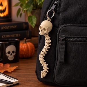 May include: A cream-colored keychain featuring a realistic human skull attached to a flexible, articulated spinal column. The keychain is attached to the zipper pull of a black fabric backpack, showcasing detailed bone textures and segments.