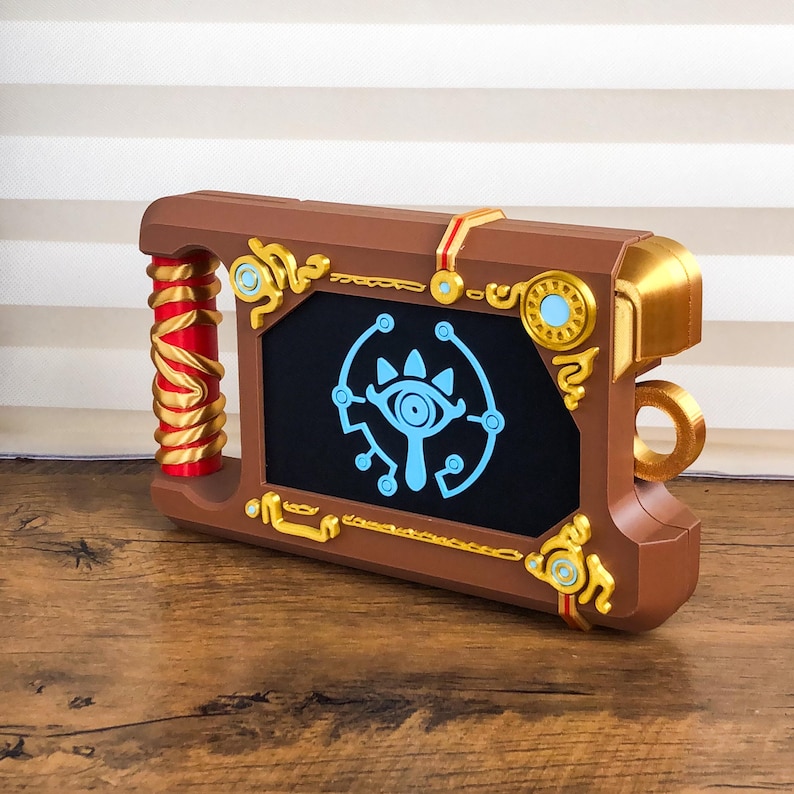 Pode incluir: Um acess&oacute;rio castanho, dourado e vermelho que se assemelha a um livro ou tablet. O dispositivo apresenta um ecr&atilde; preto com um gr&aacute;fico azul de um olho. Acentos dourados e detalhes vermelhos adornam as bordas e a pega. O acess&oacute;rio &eacute; provavelmente inspirado num videojogo.