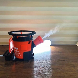 以下が含まれることがあります： 蒸気を放出する赤いノズルを備えた、オレンジと黒のデバイス。 デバイスは円形のハンドルと、側面にテキストのある長方形のボディを備えています。 デバイスは木製の表面にあります。