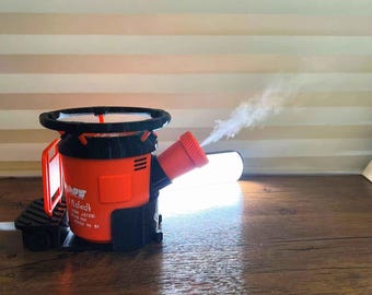 May include: An orange and black device with a red nozzle emitting a stream of vapor. The device has a circular handle and a rectangular body with text on the side. The device is on a wooden surface.