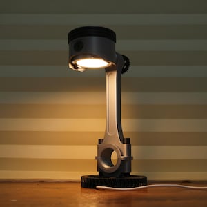 Könnte beinhalten: Eine einzigartige Schreibtischlampe aus recycelten Autoteilen. Die Lampe hat einen silberfarbenen Kolbenkopf mit eingebautem Licht, der mit einer Pleuelstange verbunden ist. Sie steht auf einem zahnradförmigen Sockel, mit einem weißen Netzkabel.