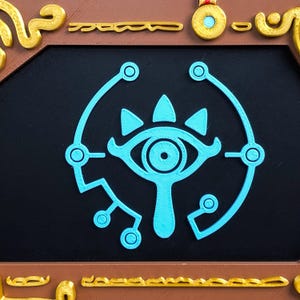 Pode incluir: Um s&iacute;mbolo de olho estilizado em azul-turquesa, provavelmente de um videogame, est&aacute; centrado em um fundo preto. O s&iacute;mbolo &eacute; cercado por uma moldura marrom e dourada. O design &eacute; provavelmente impresso em 3D.