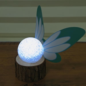 May include: A decorative LED lamp shaped like a glowing orb, set on a wooden base. The lamp is adorned with teal and white butterfly wings. The lamp emits a soft, cool light, creating a whimsical and enchanting ambiance.