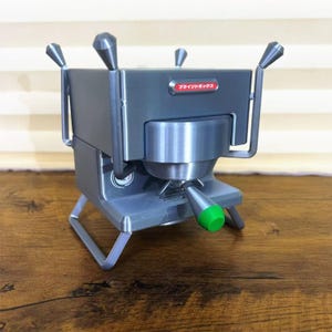 May include: A gray and silver miniature espresso machine with a green handle. The machine has a boxy design with four handles and the text "ブラインドボックス" in red. It sits on a three-legged stand.