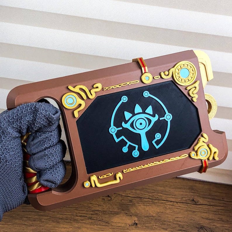 Pode incluir: Um acess&oacute;rio marrom com detalhes dourados e vermelhos, com uma tela preta com um s&iacute;mbolo de olho azul. O acess&oacute;rio tem uma al&ccedil;a e acabamentos dourados decorativos. O design lembra um videogame.