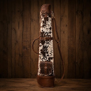 May include: A tall, slender carrying case featuring a cowhide exterior with a brown and white spotted pattern. The case includes a dark brown leather handle, a matching leather shoulder strap, and a small, rectangular leather pocket with a buckle.