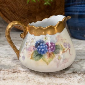 May include: A small, antique cream pitcher with a gold handle and rim. The pitcher is decorated with hand-painted blackberries in shades of blue and purple, with green leaves and white flowers. The background is a creamy white.