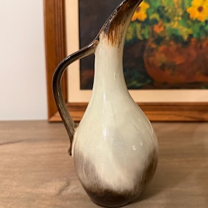 May include: A vintage ceramic pitcher with a slender, curved spout and handle. The pitcher features a gradient of colors, transitioning from a dark brown base to a creamy white body, with a brown rim and handle. The pitcher is likely used for serving beverages.