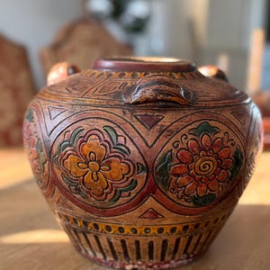 May include: A decorative, hand-carved ceramic jar with a lid. The jar features a warm brown base color with intricate floral and geometric patterns in shades of orange, red, and green. Two small handles are on the sides.