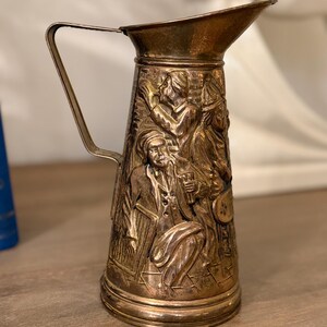 May include: A vintage metal pitcher with a detailed relief of figures in period clothing. The pitcher has a spout and a handle. The metal has a tarnished, antique appearance, suggesting age and use. The relief depicts several people in a gathering.