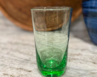 May include: A clear glass with a vibrant green base and a gradient effect. The glass has a cylindrical shape and is likely a shot glass or small drinking glass. The background includes a woven basket and a blue ceramic pot.