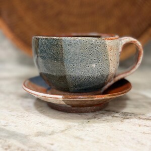 May include: A ceramic cup and saucer set featuring a faceted design with a speckled gray, blue, and dark brown glaze. The base and handle exhibit a reddish-brown clay tone, with a textured finish visible across the exterior surfaces.