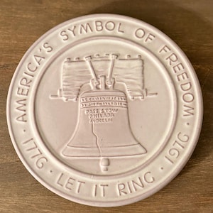 May include: A round, off-white ceramic coaster featuring the Liberty Bell. The text around the edge reads "America's Symbol of Freedom 1776 - 1976 Let It Ring". The bell is centered with the words "Pass & Stow Philadelphia" on it.