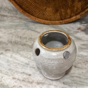 May include: A small, speckled ceramic vase with two holes. The vase is off-white with brown speckles and a brown rim. The vase has a rounded body and a pedestal base. The background includes a woven basket and a marble surface.