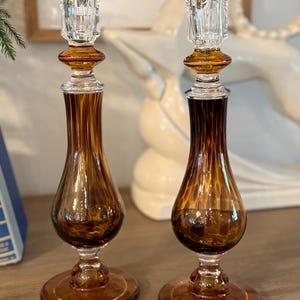 May include: Two amber-colored glass candlesticks with clear crystal tops. The candlesticks have a bulbous shape with a fluted design on the lower portion. The bases are round and the overall design is elegant and vintage.