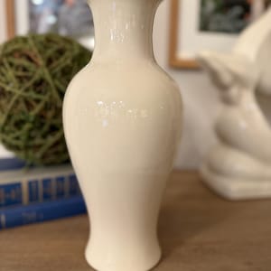 May include: A cream-colored ceramic vase with a flared top and a rounded body. The vase has a glossy finish and is displayed on a wooden surface. The vase is a decorative item, suitable for holding flowers or as a standalone accent piece.