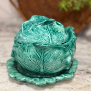 May include: A ceramic cabbage-shaped serving dish in a vibrant teal green. The dish includes a lid and a scalloped base, with detailed leaf textures. This decorative piece is ideal for kitchen decor.
