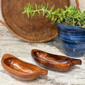 May include: Two wooden, banana-shaped bowls with a rich, warm brown finish. The bowls are smooth and curved, with a small handle-like extension at one end. They sit on a speckled, light-colored surface, with a woven basket and a blue ceramic pot in the background.