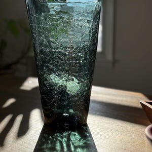 May include: A tall, teal-colored glass vase with a textured, crackled surface. The vase is cylindrical with a slightly flared top. The sunlight casts a shadow on the wooden surface below, highlighting the vase's unique texture and color.