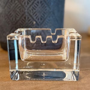 May include: A clear, square glass ashtray with a recessed center and four notched rests. The ashtray is made of thick, transparent glass, reflecting light. The design is simple and functional, suitable for home or office use.