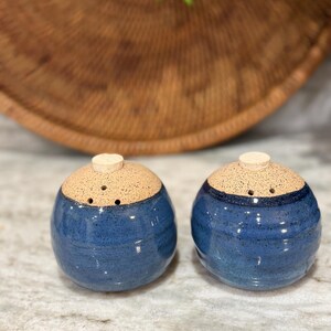 May include: A pair of ceramic salt and pepper shakers. The shakers are blue with a speckled tan top and a small wooden stopper. The shakers are round and have a glossy finish. The salt and pepper shakers are handmade.