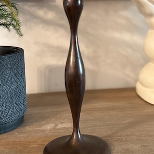 May include: A tall, dark brown wooden candlestick holder featuring a smooth, elongated, and curved silhouette. The object has a wide, circular base that tapers into a slender, hourglass-shaped stem, culminating in a cylindrical top cup.