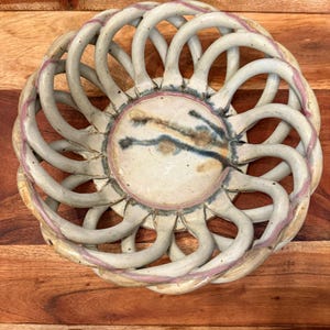 May include: A handcrafted ceramic basket with a unique openwork design. The basket features a circular shape with a central disc and a series of arched supports. The color palette includes off-white, pink, and brown tones, creating a rustic aesthetic.