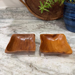 May include: Two small, square wooden bowls with a warm, reddish-brown finish. The bowls have a slightly irregular, organic shape, and are displayed side-by-side. The wood grain is visible, adding to their natural aesthetic. These bowls are suitable for serving snacks or small items.