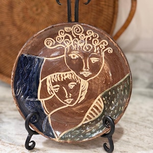 May include: A round ceramic plate featuring a sgraffito design of two faces in brown clay. The left side has a dark blue textured section, while the right side features a green dotted pattern. The plate is displayed on a black metal stand.