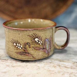 May include: A tan ceramic mug with a brown rim and handle. The mug is decorated with wheat stalks in shades of brown, white, and purple. The mug is sitting on a light-colored surface.
