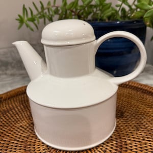 May include: A white ceramic teapot with a lid, spout, and a rounded handle. The teapot is sitting on a woven wicker tray. Green plants are visible in the background.