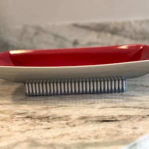 May include: A rectangular ceramic serving dish with a red interior and a gray exterior. The dish is supported by a rectangular base with vertical blue and white stripes. The dish is on a marble countertop.