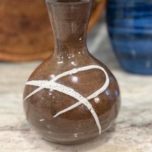 May include: A small, round, brown ceramic vase with a narrow neck and a flared rim. The surface features a glossy finish with a hand-painted, white, speckled, abstract line design that curves across the front of the rounded body of the vessel.