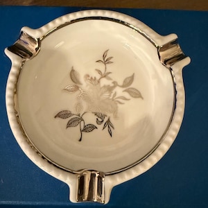 May include: A vintage white ashtray with a gold rim and accents. The ashtray features a floral design in silver and gold tones. It has four rests for cigarettes and a scalloped edge.