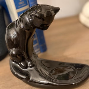 May include: A glossy black ceramic cat figurine with a curved body and a small dish. The cat is in a seated position, looking down. The dish is oval-shaped. The figurine is a decorative item.