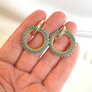 Puede incluir: Pendientes de aro dorados con diseño de cuentas. Los pendientes presentan una combinación de colores turquesa y dorado, con las cuentas dispuestas en un patrón circular. Los aros se sujetan con un cierre dorado.