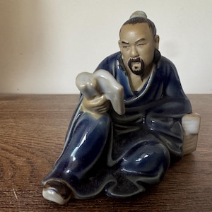 Op de afbeelding: Een keramieken beeldje van een zittende Aziatische man in een donkerblauwe jurk. Het beeldje heeft een gedetailleerd gezicht met een baard en houdt een lichtgekleurd voorwerp vast. Het beeldje staat op een houten oppervlak.