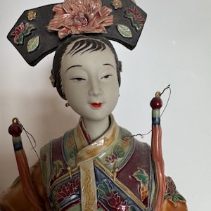 May include: A porcelain figurine of a woman in traditional Chinese attire. The figurine features a detailed floral headdress, a colorful robe with intricate patterns, and two decorative tassels. The face has delicate features and red lips.