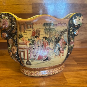 Può includere: Un vaso antico in ceramica con una scena asiatica dipinta a mano. Il vaso presenta un corpo color crema con una dettagliata raffigurazione di figure in abiti tradizionali, ambientata in un paesaggio. Le maniglie e i bordi sono neri con decorazioni floreali.