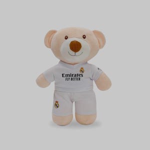 以下が含まれることがあります： 白いサッカーユニフォームを着たテディベア。ユニフォームには「Emirates FLY BETTER」の文字とチームのロゴが入っています。クマは薄茶色の鼻をしていて、笑顔です。この衣装には、ジャージとショーツが含まれています。