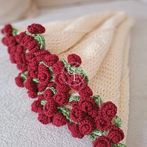 Puede incluir: Manta de ganchillo color crema con un borde de rosas rojas de ganchillo y hojas verdes. La manta está doblada, mostrando los adornos florales a lo largo del borde. Las rosas están densamente empaquetadas, creando un ribete decorativo texturizado.