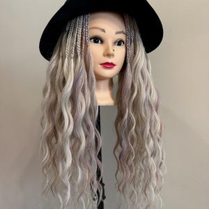 Könnte beinhalten: Ein Schaufensterpuppenkopf mit langem, welligem, hell gefärbtem Haar, das mit Zöpfen und einem schwarzen Hut gestylt ist. Das Haar hat subtile rosa und blaue Highlights. Die Schaufensterpuppe hat einen neutralen Ausdruck mit rotem Lippenstift.