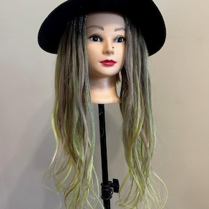 Puede incluir: Una cabeza de maniquí con cabello largo y multicolor en tonos verdes, marrones y rubios, peinado con trenzas en la coronilla. El maniquí lleva un sombrero de fieltro negro de ala ancha. La cabeza está montada sobre un soporte negro.