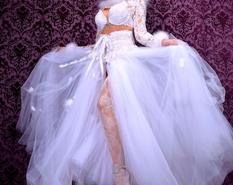 May include: A woman wearing a white lace dress with a tulle skirt and a white fur trim. She is wearing a silver tiara and white lace stockings. The background is a purple patterned wall.